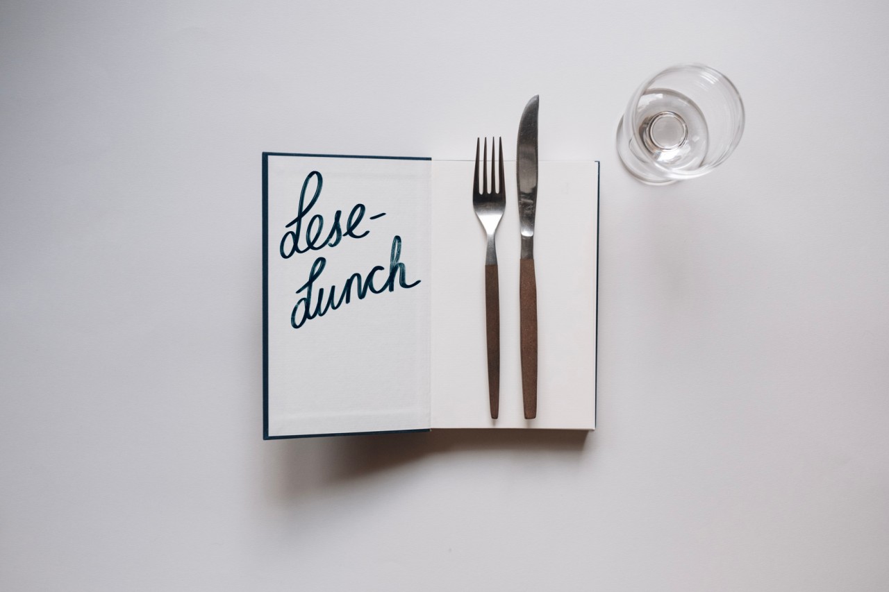Auf einem weissen Untergrund liegt ein aufgeschlagenes Buch. Auf der Innenseite des Umschlags steht Lese-Lunch geschrieben, auf der rechten Seite liegen eine Gabel und ein Messer. Rechts oben neben dem Buch steht ein leeres Weinglas.