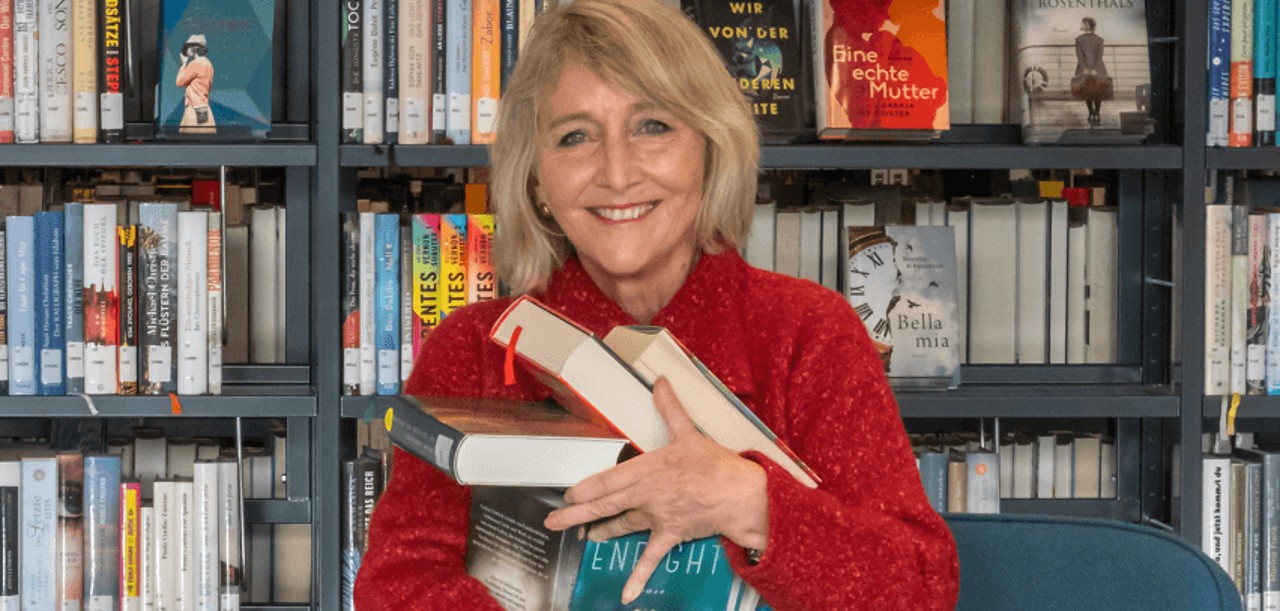 Frau mit rotem Pullover vor einem Bücherregal mit mehreren Büchern im Arm.