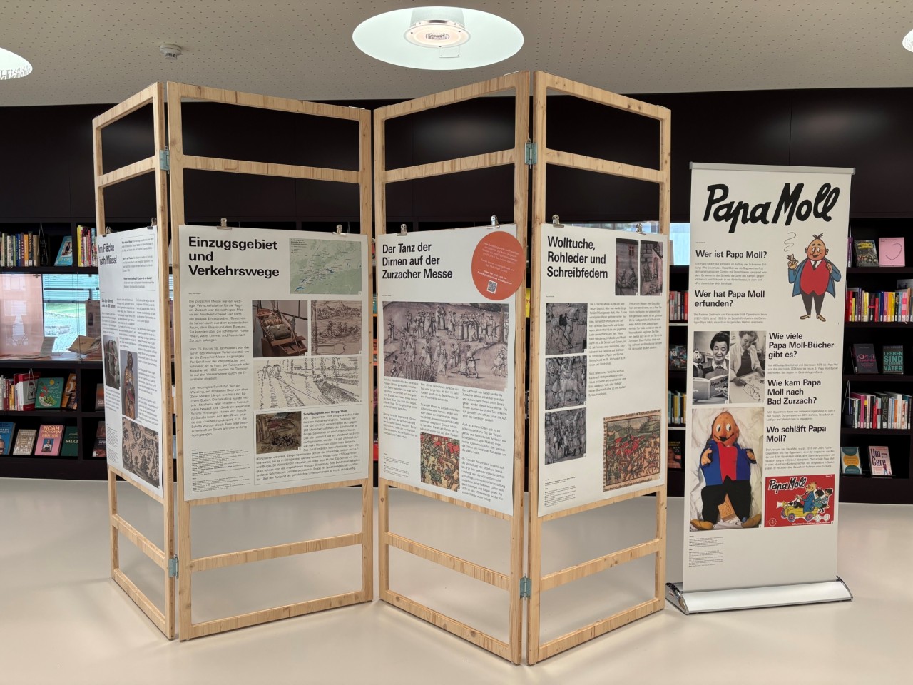 Plakate an Stellwänden aus Holz vor einem Bücherregal. Rechts daneben ein Rollup.