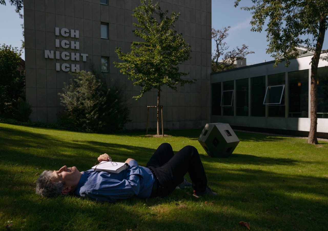 Ein Mann liegt auf einer grünen Wiese vor einem Gebäude mit der Schrift "ICH NICHT ICH". Im Hintergrund sind ein Baum und moderne Architektur erkennbar.