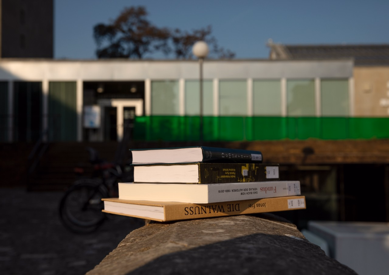 Bücherstapel auf einem Stein in der Morgensonne. Im Hintergrund die Aargauer Kantonsbibliothek mit einem grünen Plexiglasgeländer und Glasfassade.