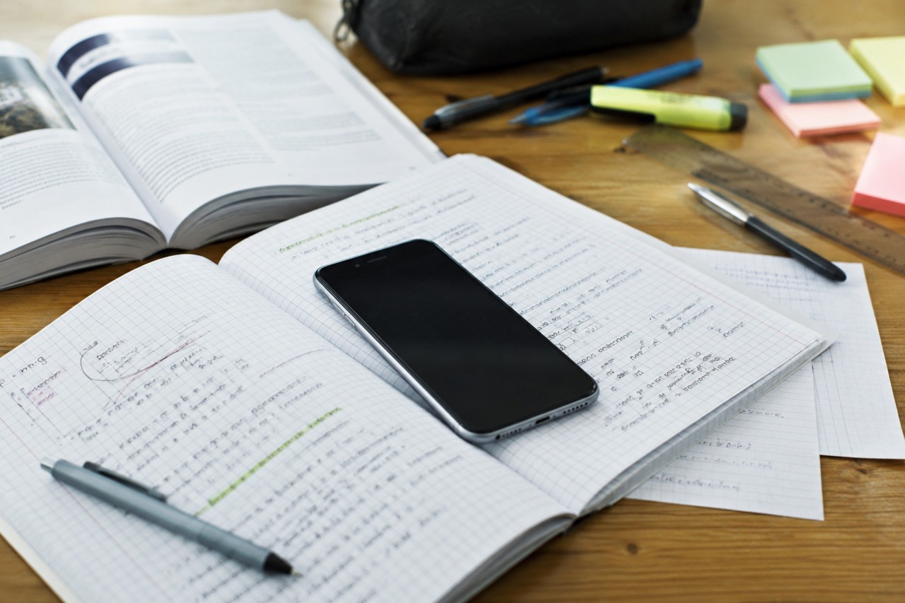 Schreibutensilien und Lernmaterialien auf einem Holztisch, darunter offene Bücher, ein Notizheft mit mathematischen Formeln, ein Smartphone, ein Lineal, Stifte, Textmarker und Haftnotizen.