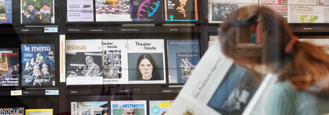 Blick durch Glas auf eine Frau, die vor einem Bücherregal sitzt und eine Zeitung liest.