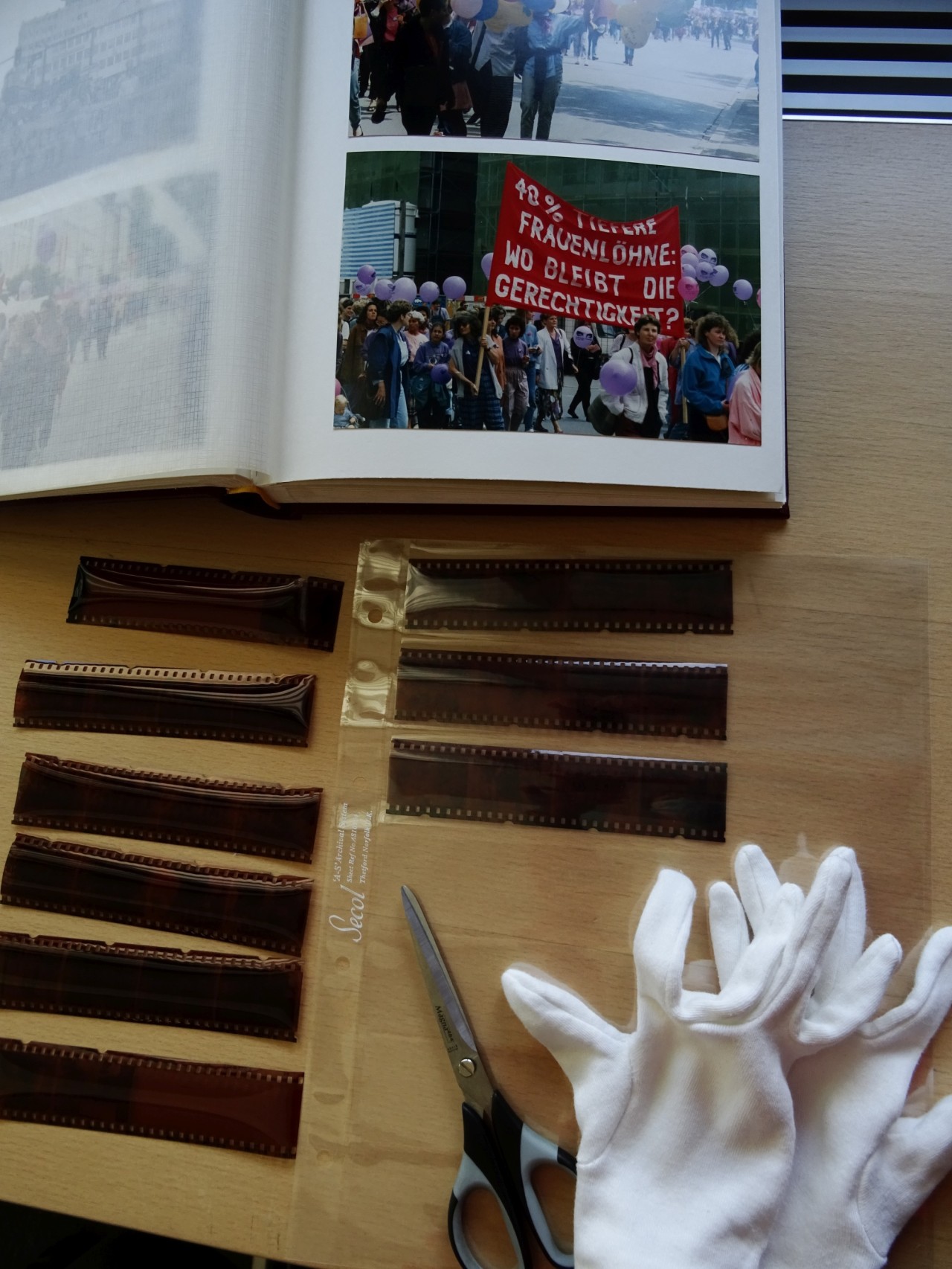 Ein aufgeschlagenes Buch mit Fotos, mehrere Filmstreifen, eine weisse Stoffhandschuhpaar und eine Schere auf einem Holztisch.