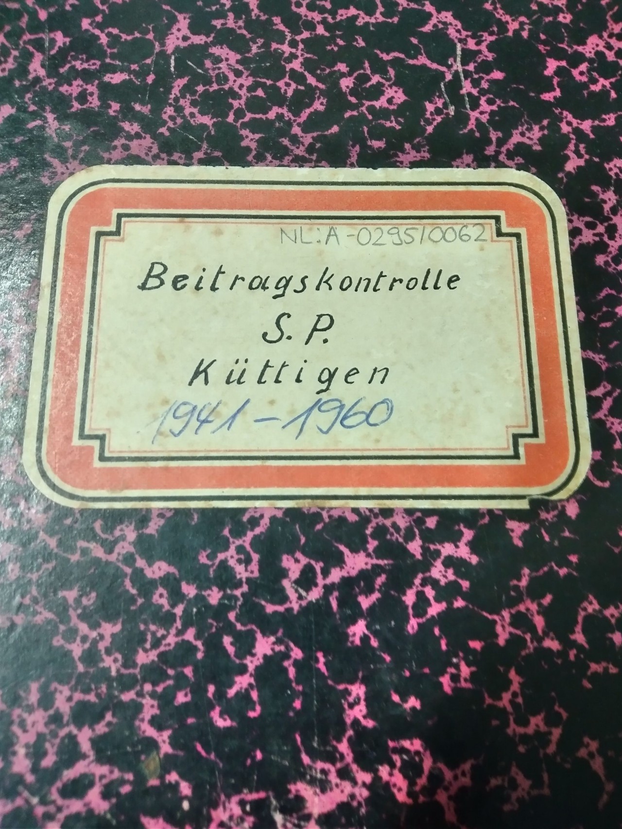 Nahaufnahme eines roten Etiketts mit goldener Umrandung auf einem schwarzen Buchdeckel mit pinkfarbenen Sprenkeln. Auf dem Etikett steht handschriftlich: Beitragskontrolle, S.P., Küttigen, 1941–1960.