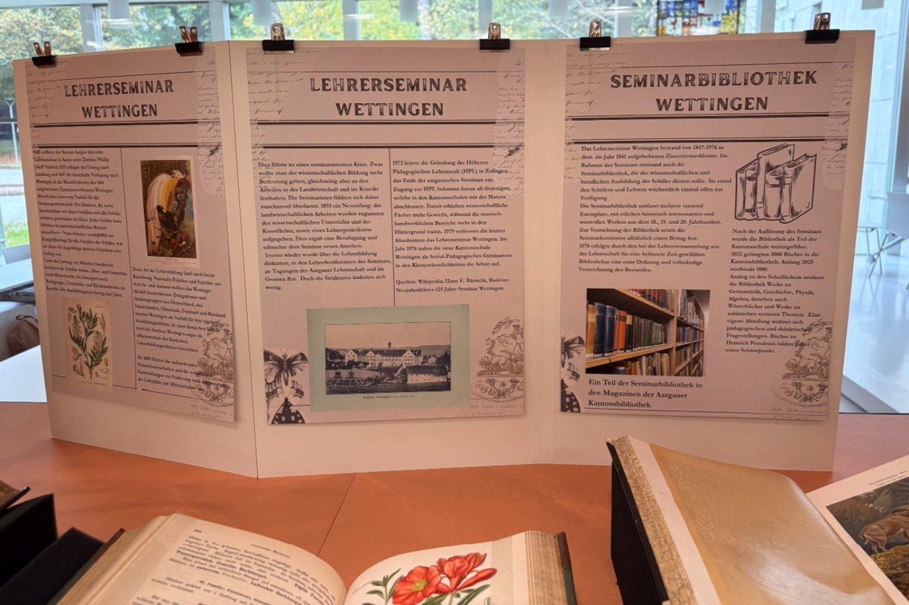 Blick in eine geöffnete Vitrine auf gestaltete Stellwand mit Text und Bild und aufgeschlagene, farbig illustrierte Bücher.