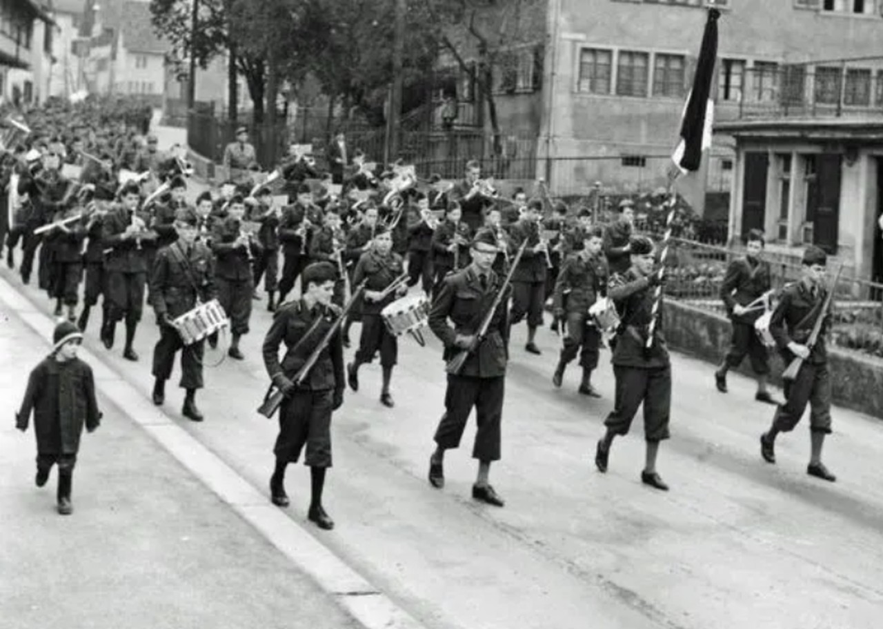 Schwarz-weiss-Foto zeigt einen Kadettenumzug von jungen Männern in Uniformen mit Musikinstrumenten und Fahnen.
