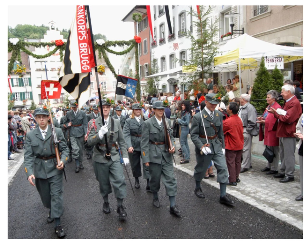 Kadettenzug mit jungen Männern in Uniform und Fahnen durch die geschmückte Altstadt. Zuschauende links und rechts der Strasse.