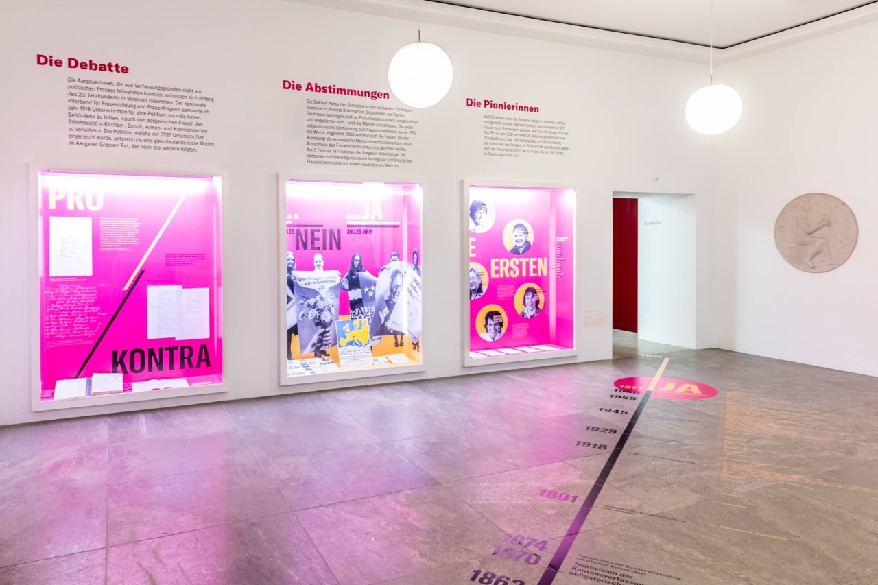 Raum mit drei Glasvitrinen mit Text und Bild zum Frauenstimmrecht auf pinkem Hinetrgrund.