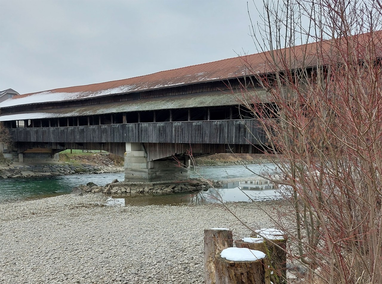 Foto einer Holzbrücke über einem Fluss mit Kiesbett.