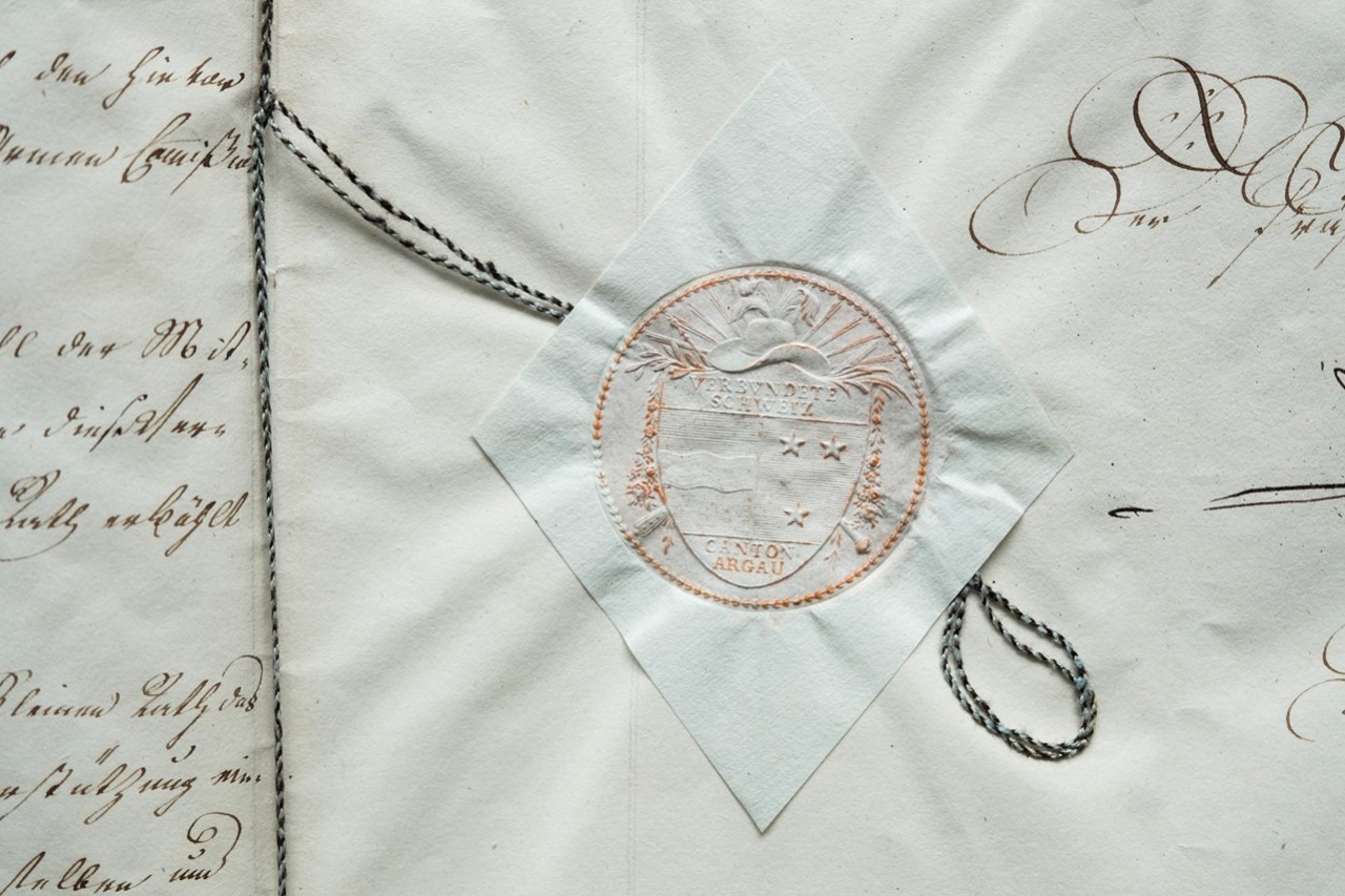 Das Bild zeigt ein Detail eines Siegels beziehungsweise eines Blindprägung eines Siegels aus dem Kanton Aargau. Das Detail schmückt das Armengesetz zum Schluss des Dokuments.