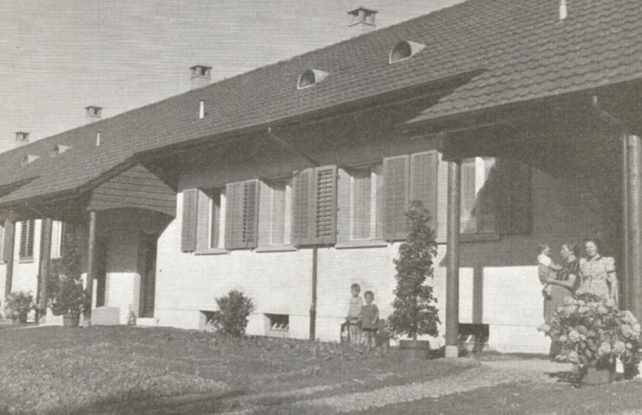 Schwarzweiss-Fotografie. Zeigt die der Strasse zugewandten Hauseingänge. Hauseingänge sind überdacht. Frauen und Kinder sind sichtbar. Auf dem Dach hat es für jedes Haus einen Kamin (Holzheizung).