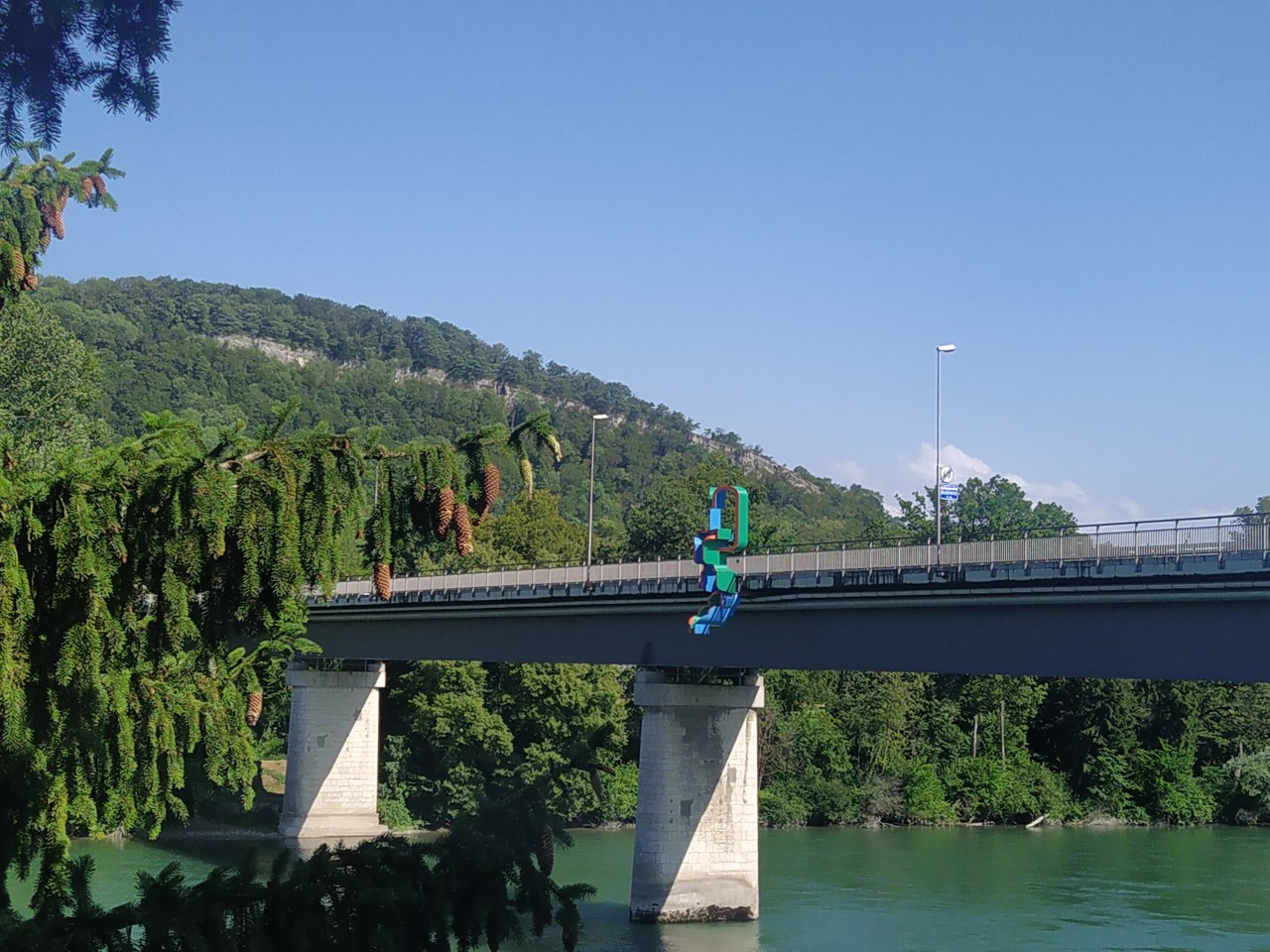 Farbfoto einer Autobrücke über einen Fluss. Am Geländer eine Kunstskulptur.