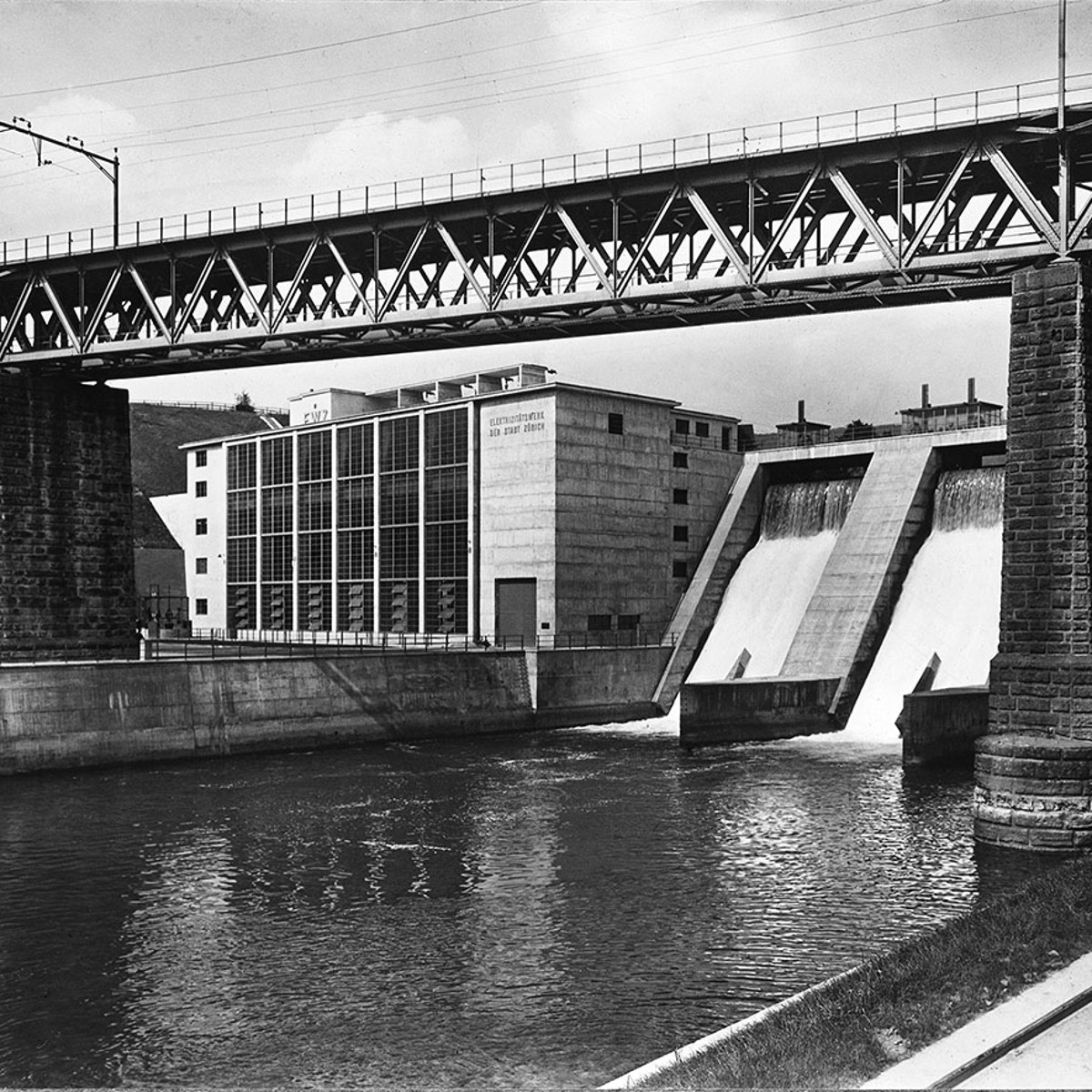 Eine schwarz-weiss Fotografie des ewz Wettingen mit Blick auf die Staumauer und die Bahngleise, die oben drüber ziehen.