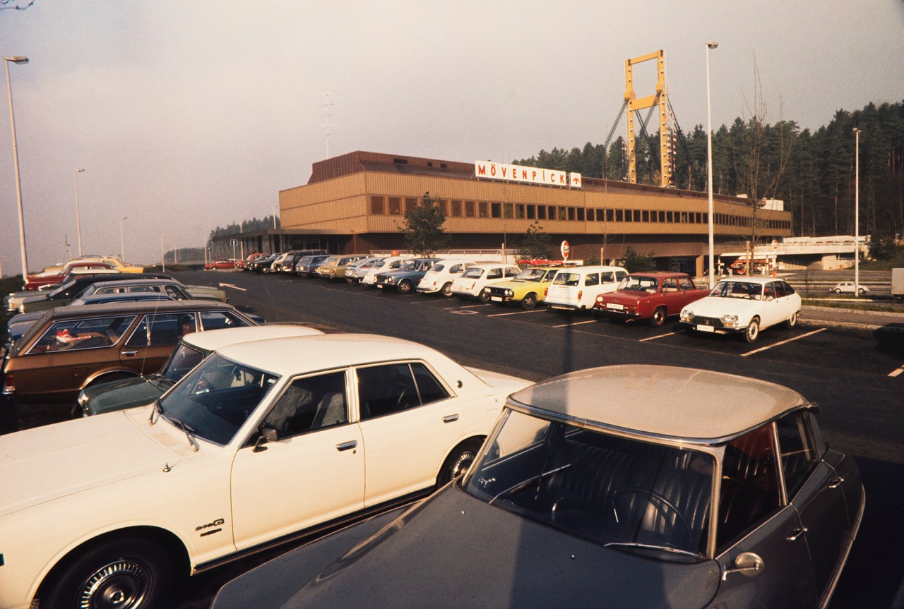 Alte Farbaufnahme einer Brücke mit Mövenpick-Restaurant. Im Vordergrund ein Parkplatz mit Autos von früher.