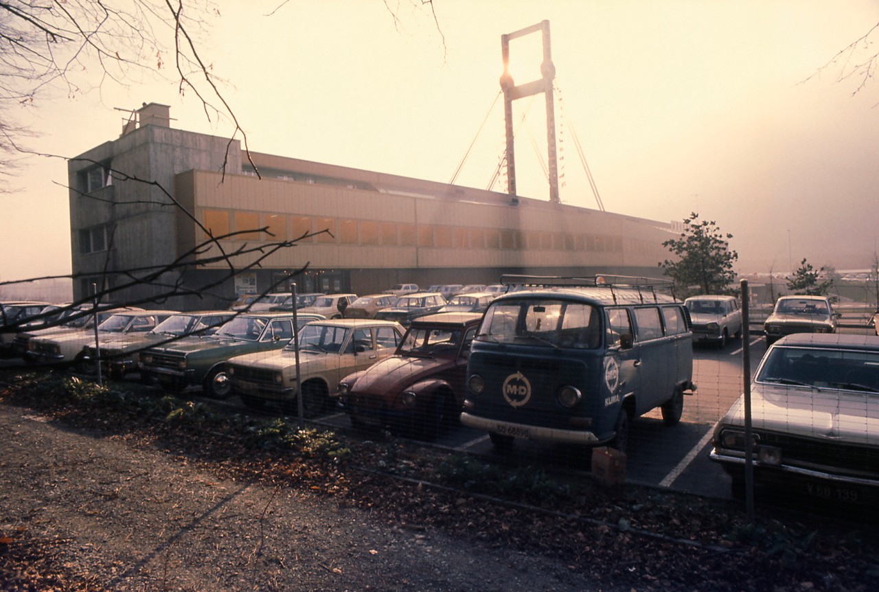 Alte Farbaufnahme einer Brücke mit Autobahnraststätte und Parkplatz mit Autos.