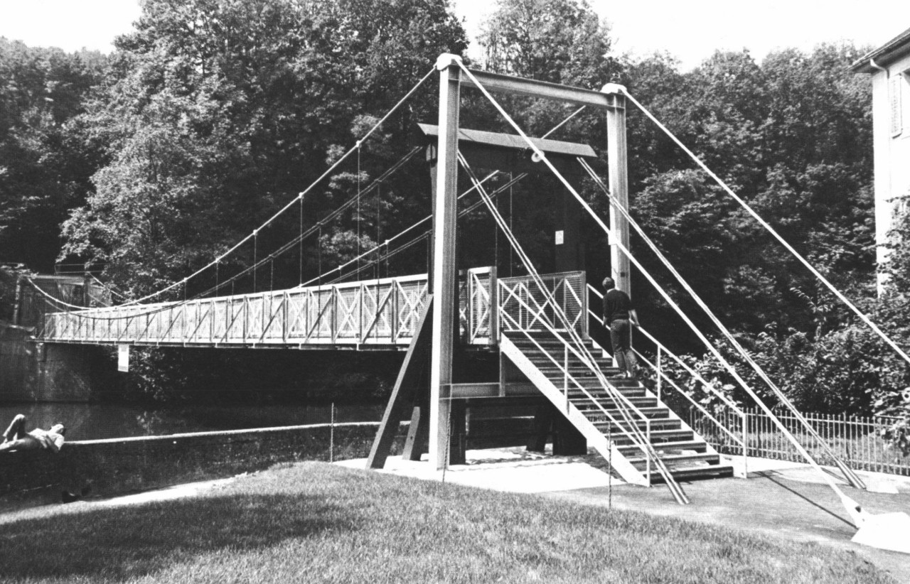 Schwarz-weiss-Foto einer Hängebrücke über einen Fluss mit Treppe.