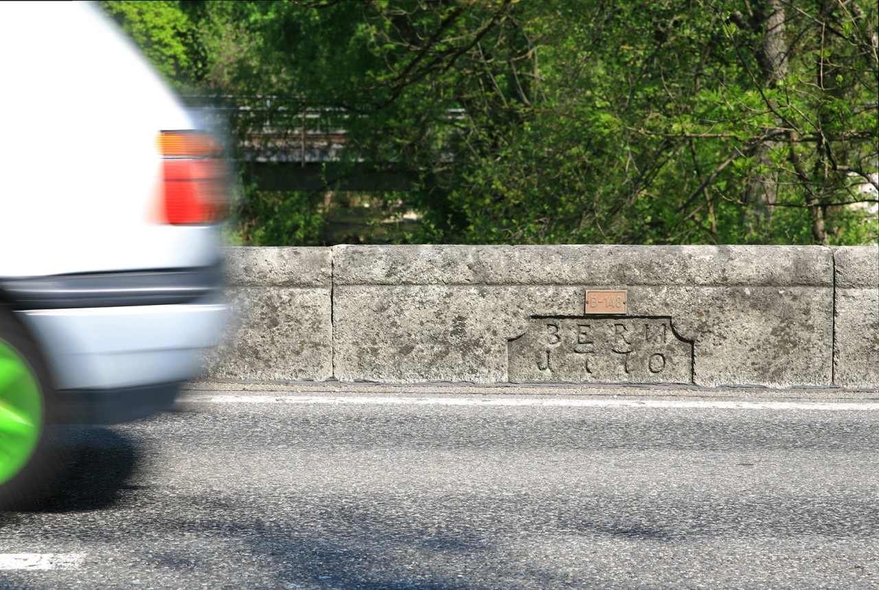 Farbfoto auf dem ein wegen der Fahrt leicht verschwommenes Heck eines Autos mit grünen Felgen zu erkennen ist. Vermutlich auf einer Brücke aus Stein mit Baujahr an der Mauer vermerkt. Dahinter Bäume.