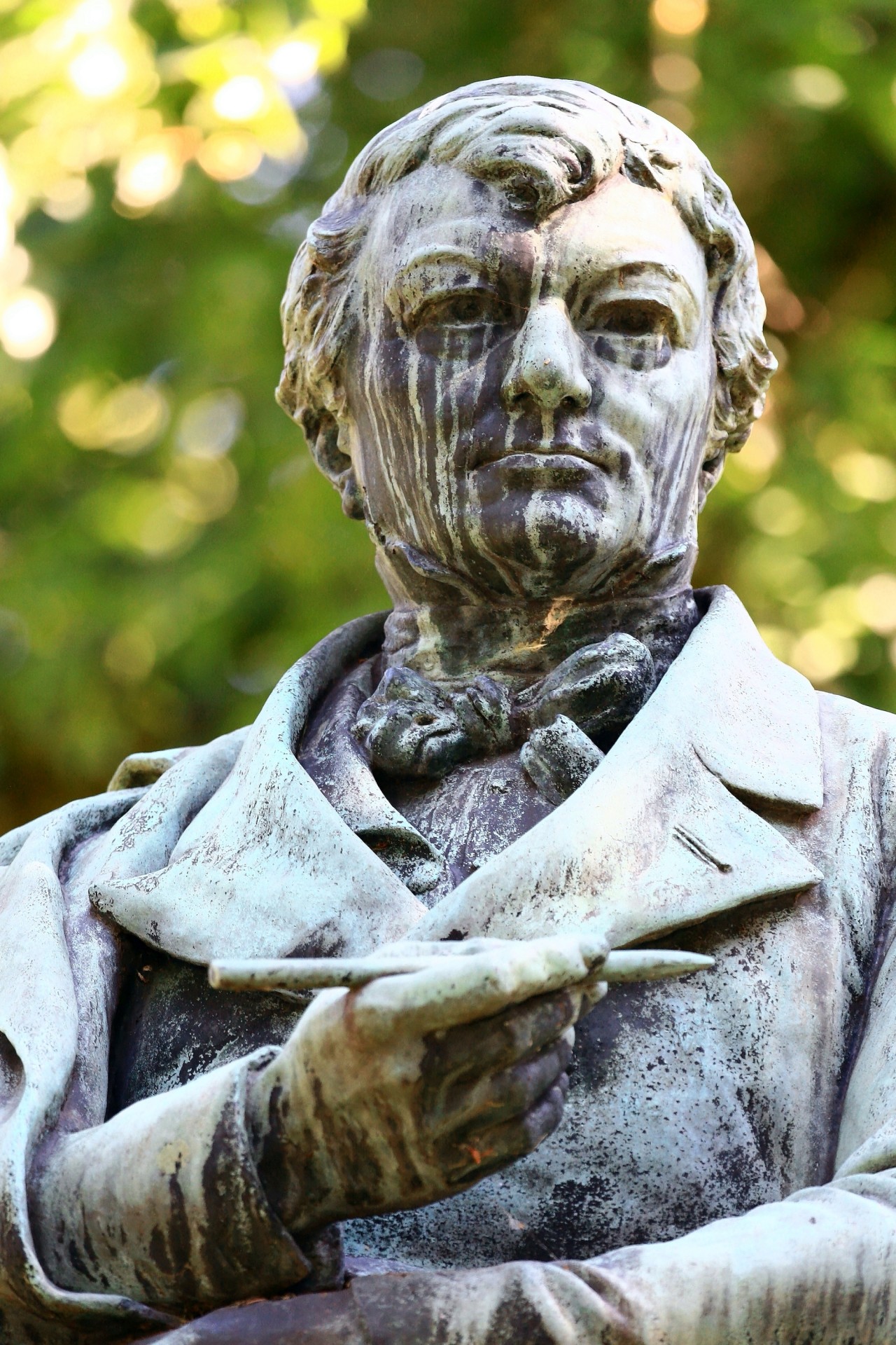 Statue eines Mannes mit Anzug und Fliege, der eine Schriftrolle in der rechten Hand hält, mit grünem Hintergrund aus unscharfen Bäumen.