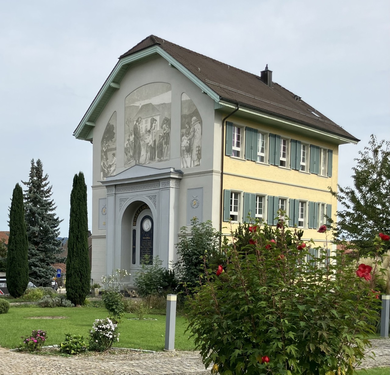 Schulhaus mit Rosenstrauch, grosser Malerei an der Fassade und Gedenktafel.