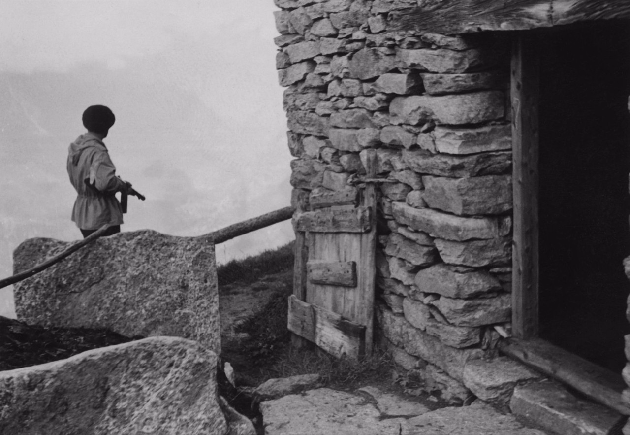 Schwarz-weiss-Foto zeigt einen bewaffneten Mann neben einem Steinhaus.