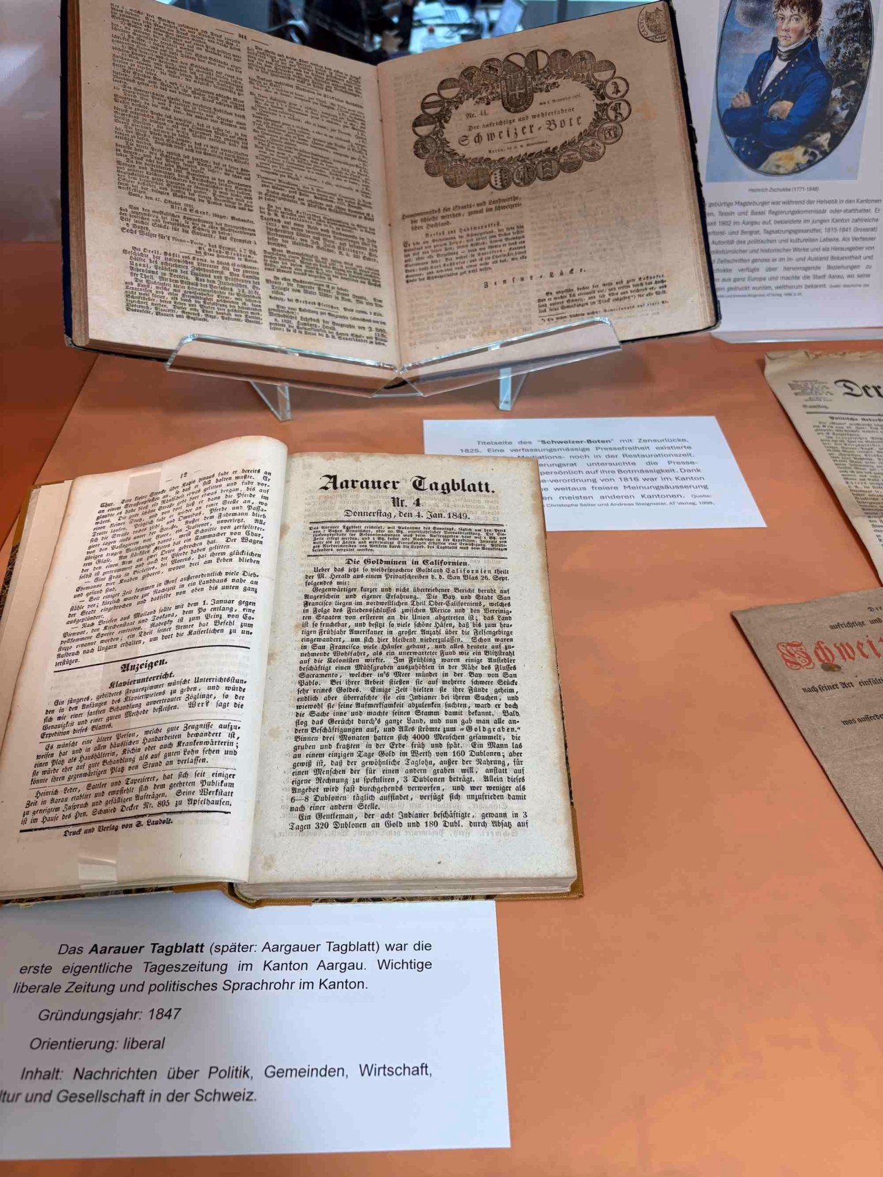 Aufgeschlagene Zeitungsbände in einer Vitrine. Davor jeweils Texte auf weissem Papier.