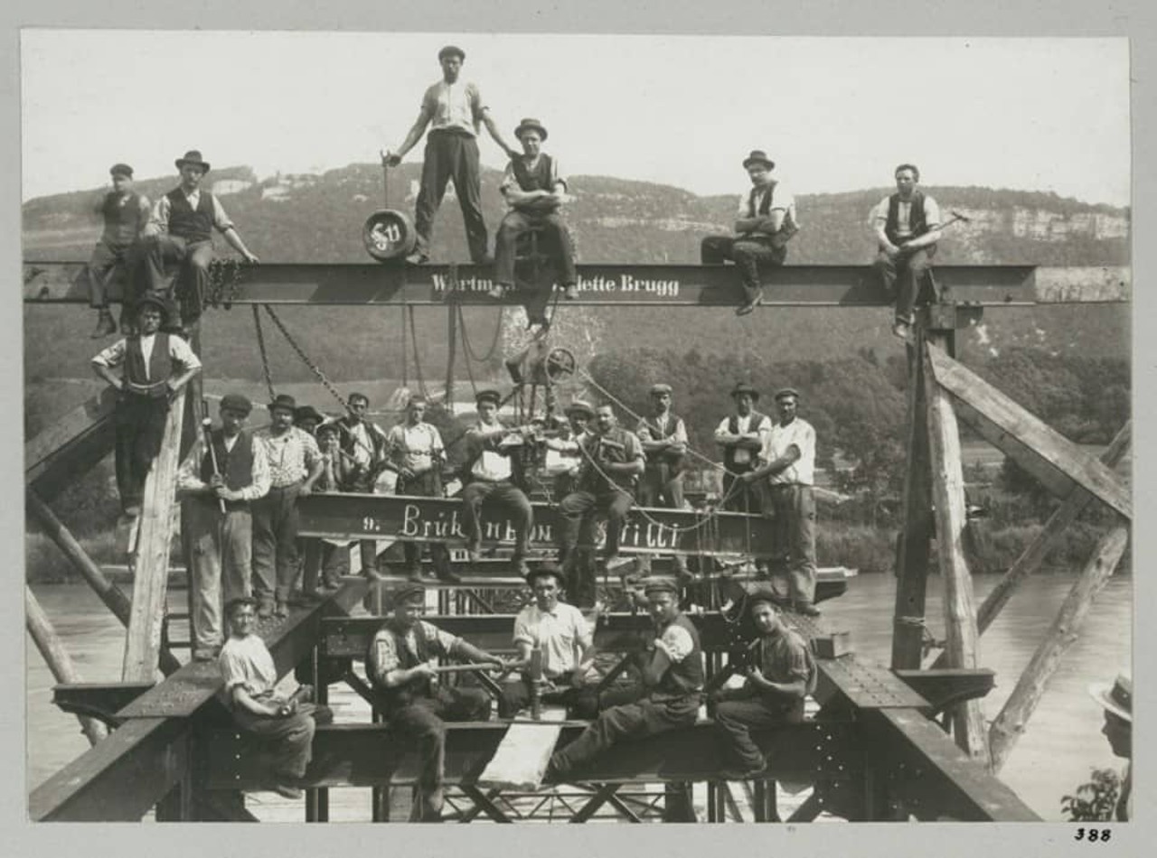 Schwarz-weiss-Foto von Arbeitern sitzend und stehend auf einer Brückenkonstruktion.