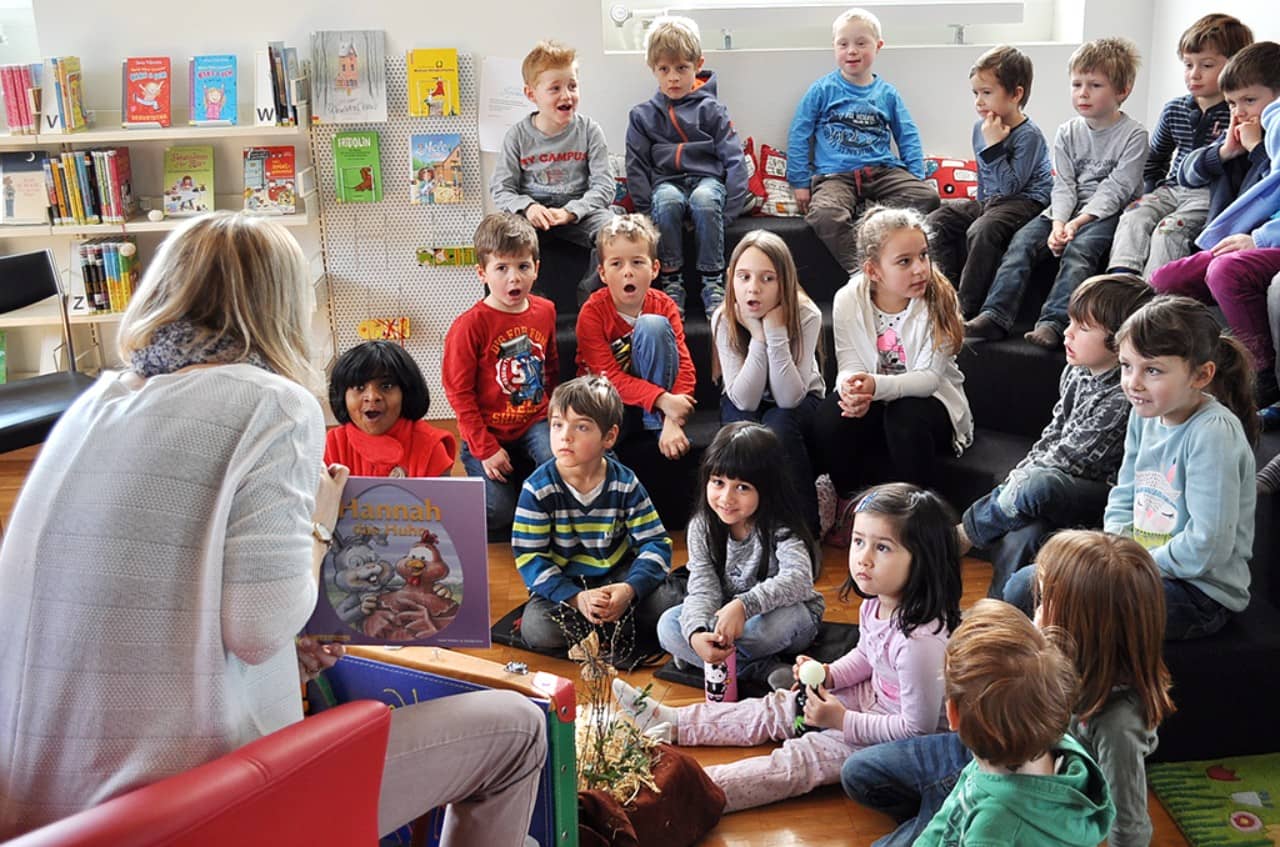 Eine Frau zeigt ein Bilderbuch, eine Gruppe Kinder ihr gegenüber lauscht und staunt