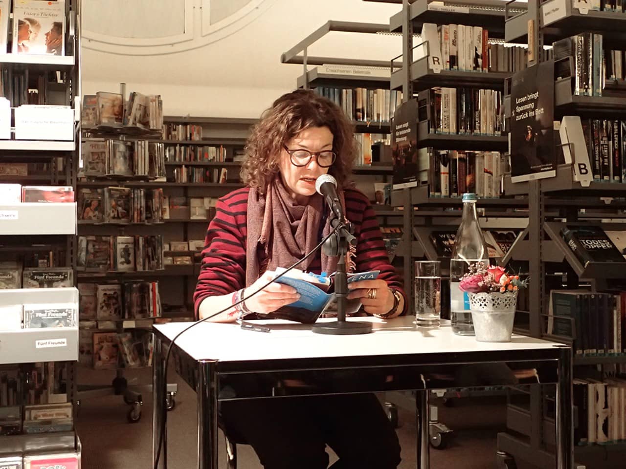 Milena Moser sitzt an einem Tisch zwischen Bücherregalen und liest aus ihrem Buch.
