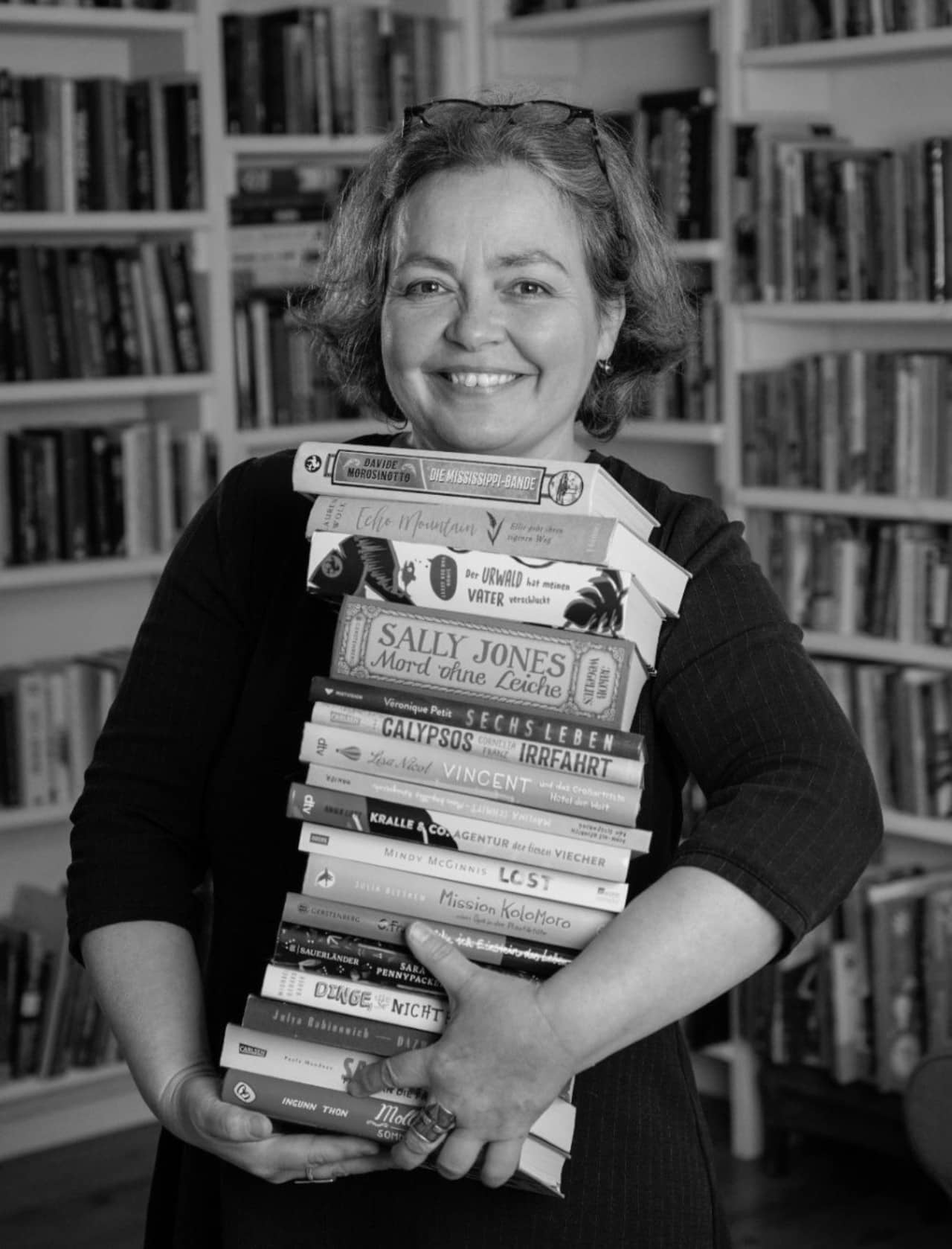 Schwarz-weiss Porträt von Tina Kemnitz mit Bücherstappel in den Händen vor Bücherregalen.