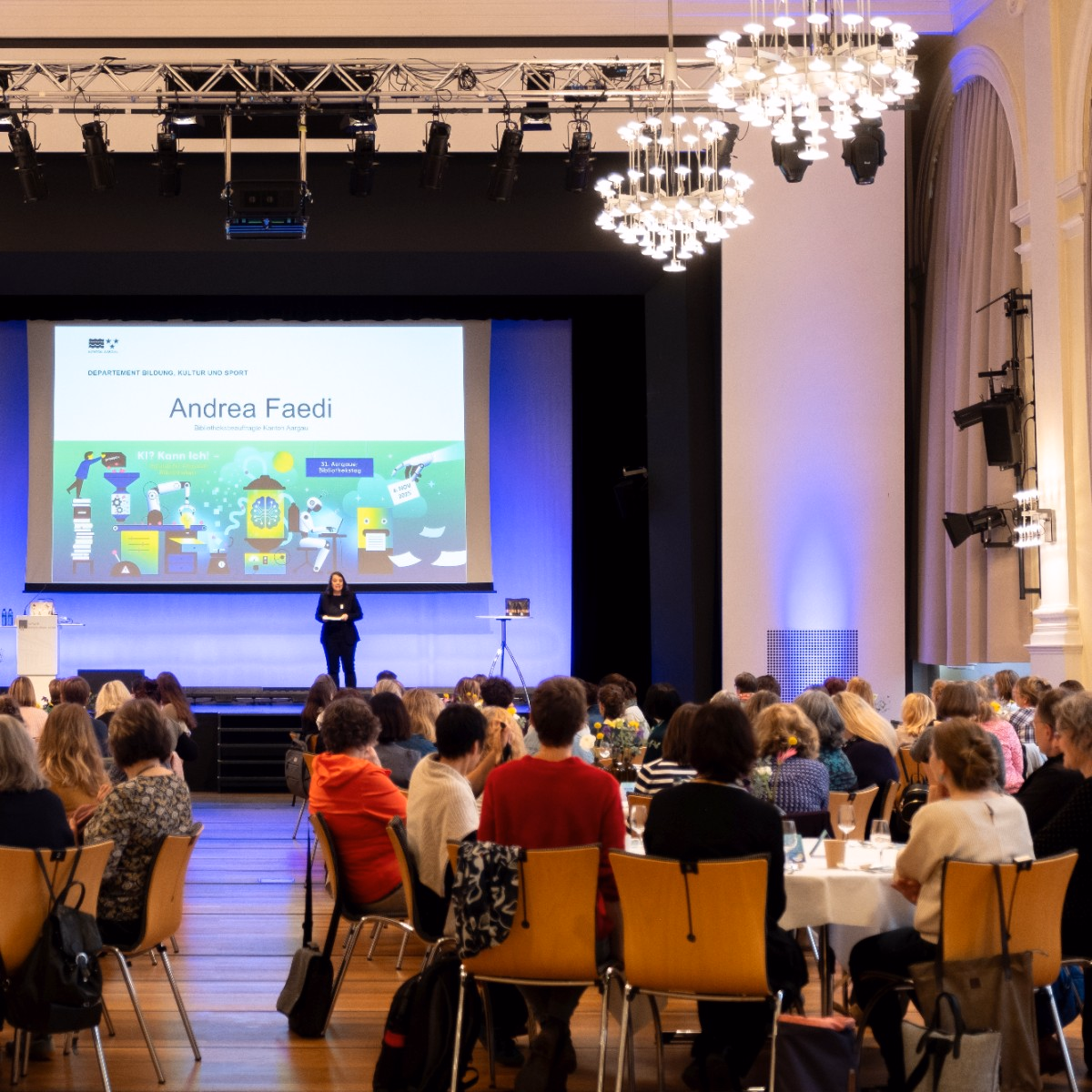 Im Saal mit Kronleuchtern sitzen Personen an runden Tischen und schauen zur Bühne. Auf der Bühne spricht eine Frau. Im Hintergrund ist eine Präsentation projiziert.