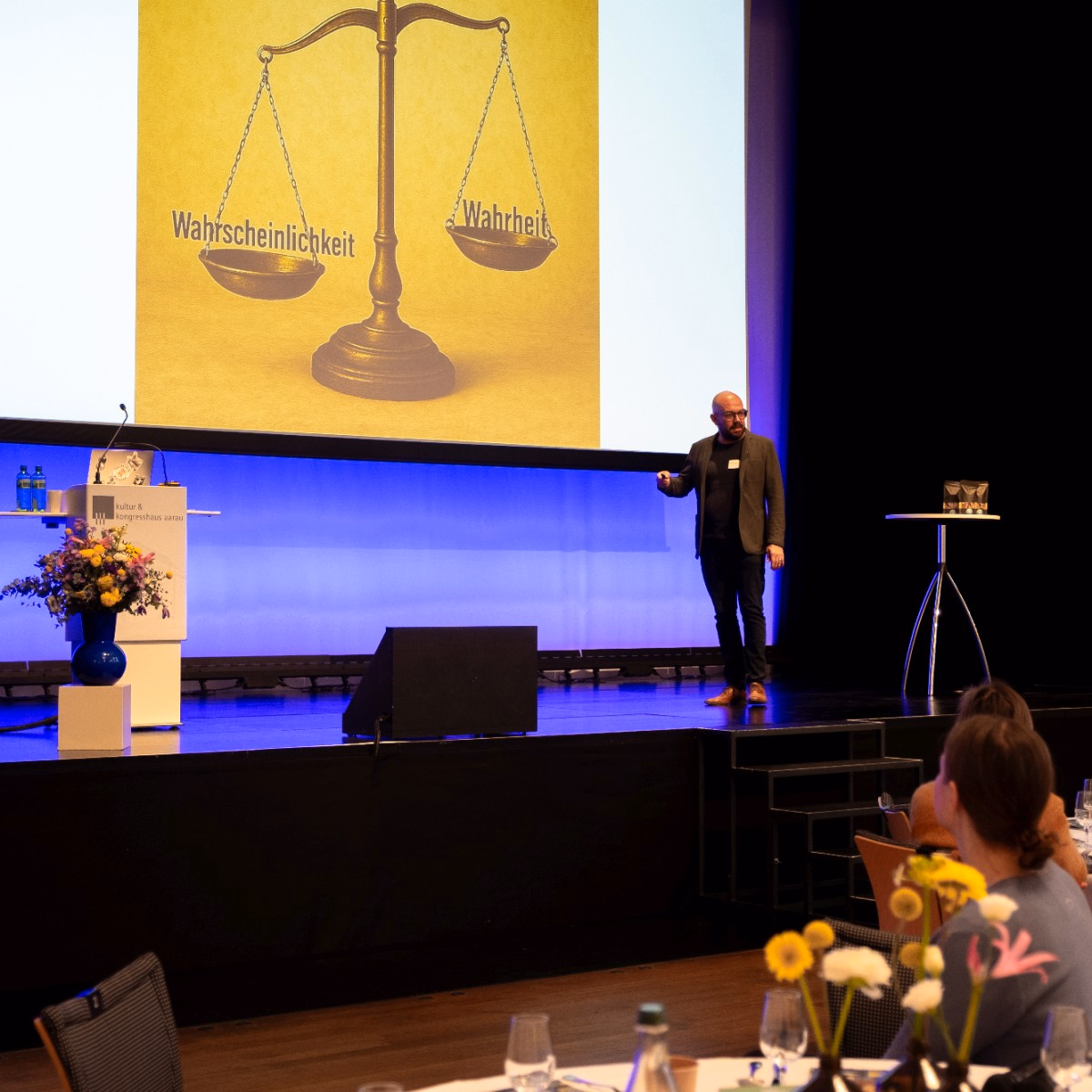 Mann präsentiert auf einer Bühne eine Präsentationsfolie mit einer Waage mit den Begriffen Wahrheit und Wahrscheinlichkeit.