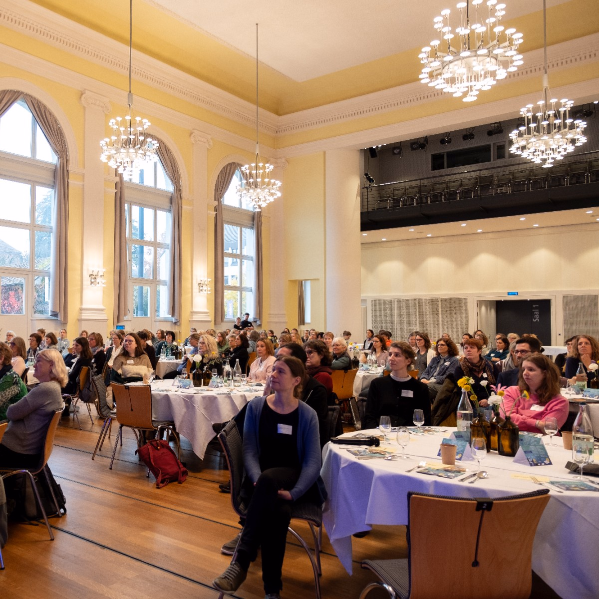 Publikum in einem Saal mit Kronleuchtern an runden Tischen.