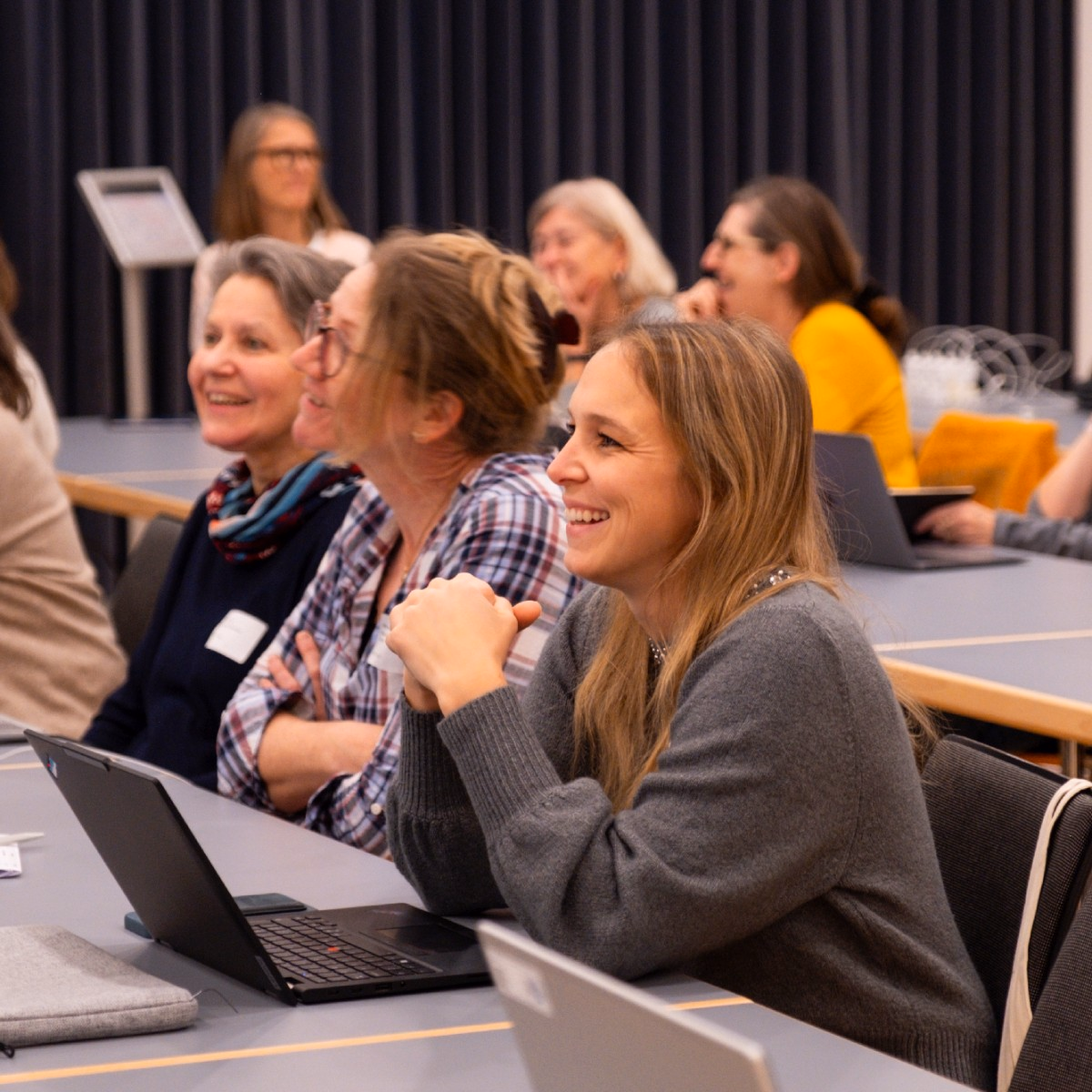 Frauen sitzen nebeneinander mit Laptop an Tischen und lachen.