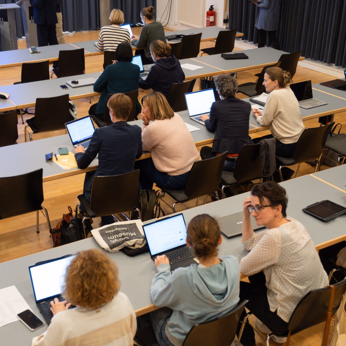 Personen sitzen mit Laptops in einem Sitzungszimmer an Tischen und diskutieren miteinander.