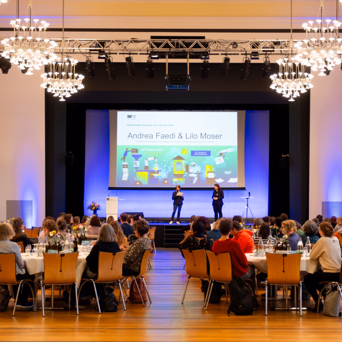 Im Saal mit Kronleuchtern sitzen Personen an runden Tischen und schauen zur Bühne. Auf der Bühne spricht eine Frau. Im Hintergrund ist eine Präsentation projiziert.