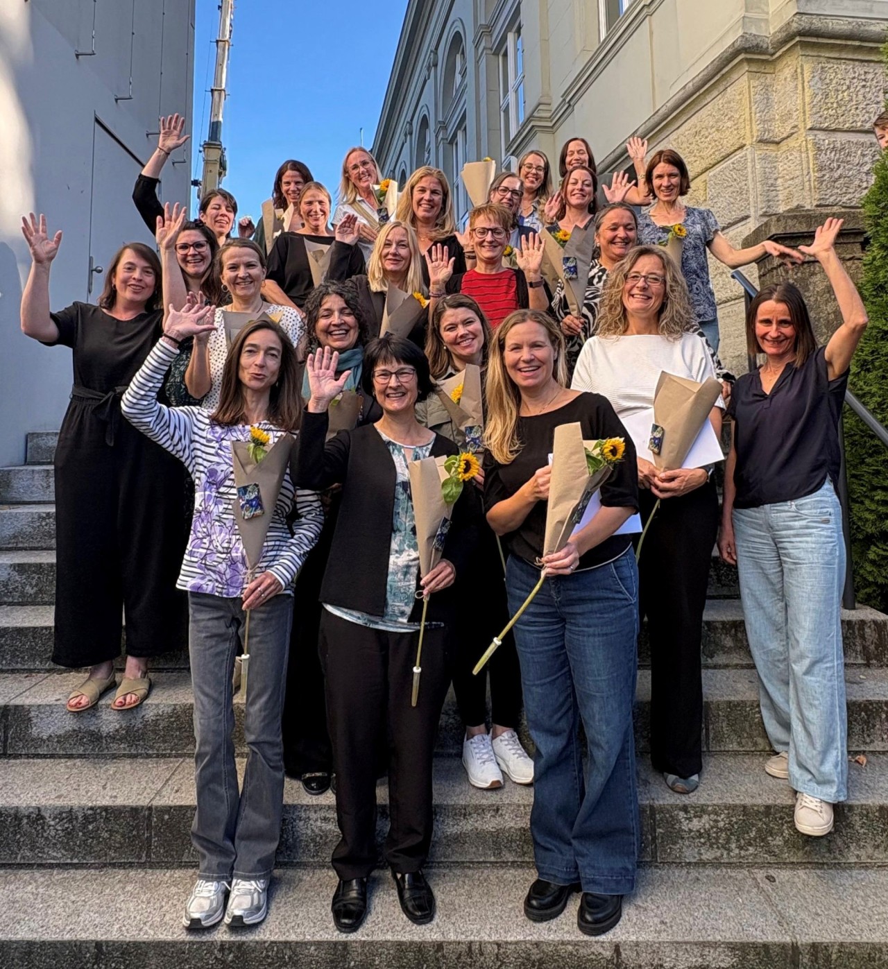 Gruppe von rund 25 Frauen, die auf einer Treppe vor einem Gebäude stehen, Blumensträusse halten und freundlich in die Kamera winken.