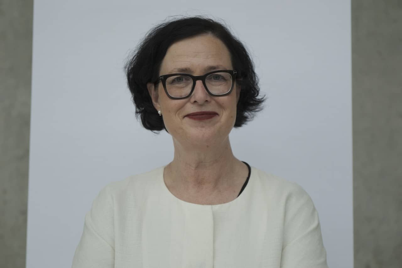 Das Portrait zeigt eine Frau vor einer Wand von Kopf- bis Bauchhöhe. Sie trägt kinnlanges, dunkles, gewelltes Haar, eine schwarze Korrekturbrille und einen Lippenstift in Dunkelrot. Sie lächelt sanft in die Kamera.