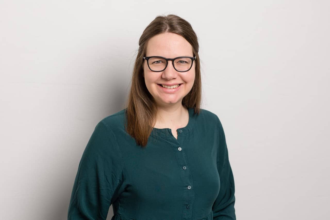 Ein Portrait von Melanie Müller. Sie steht vor einer weissen Wand und trägt ein dunkelgrünes Oberteil und Brille. Sie lächelt in die Kamera.