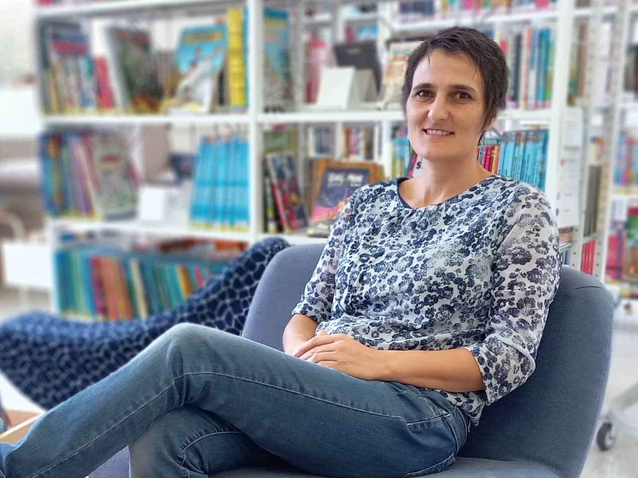 Eine Frau im mittleren Alter und kurzen, braunen Haaren sitzt entspannt auf einem Sessel in einer Bibliothek und lächelt in die Kamera. Im Hintergrund sind unscharf die Bücherregale zu erkennen.