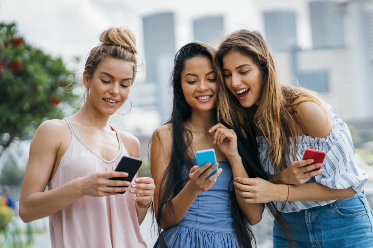 Werbefoto von drei jungen Frauen, die gemeinsam in ihre Handys schauen und begeistert lachen.