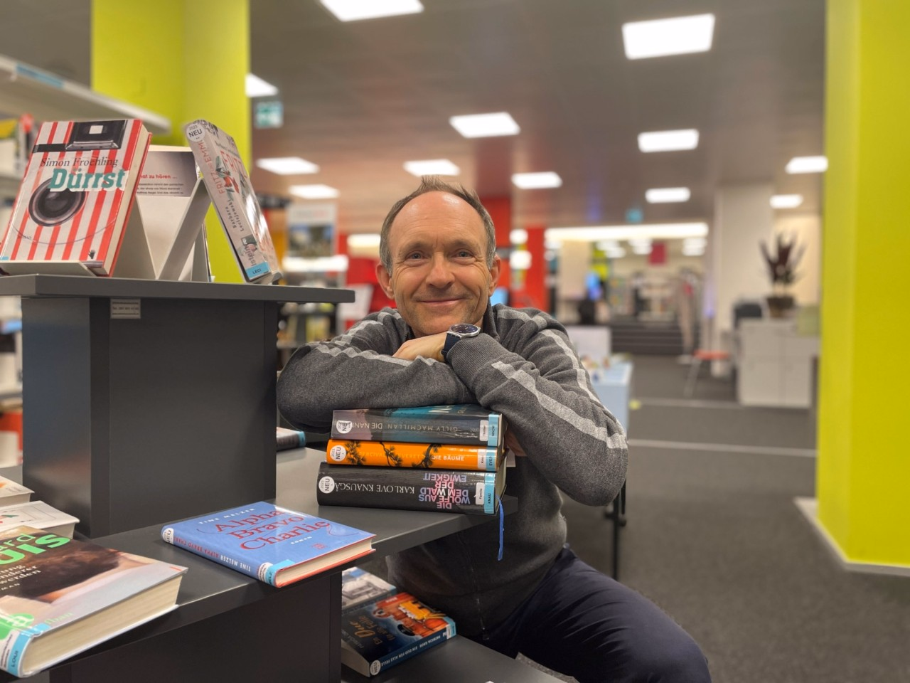 Porträt eines lächelnden Mannes mit grauem Pullover, der in einer Bibliothek neben Büchern sitzt und die Arme auf einen Stapfel Bücher stützt.