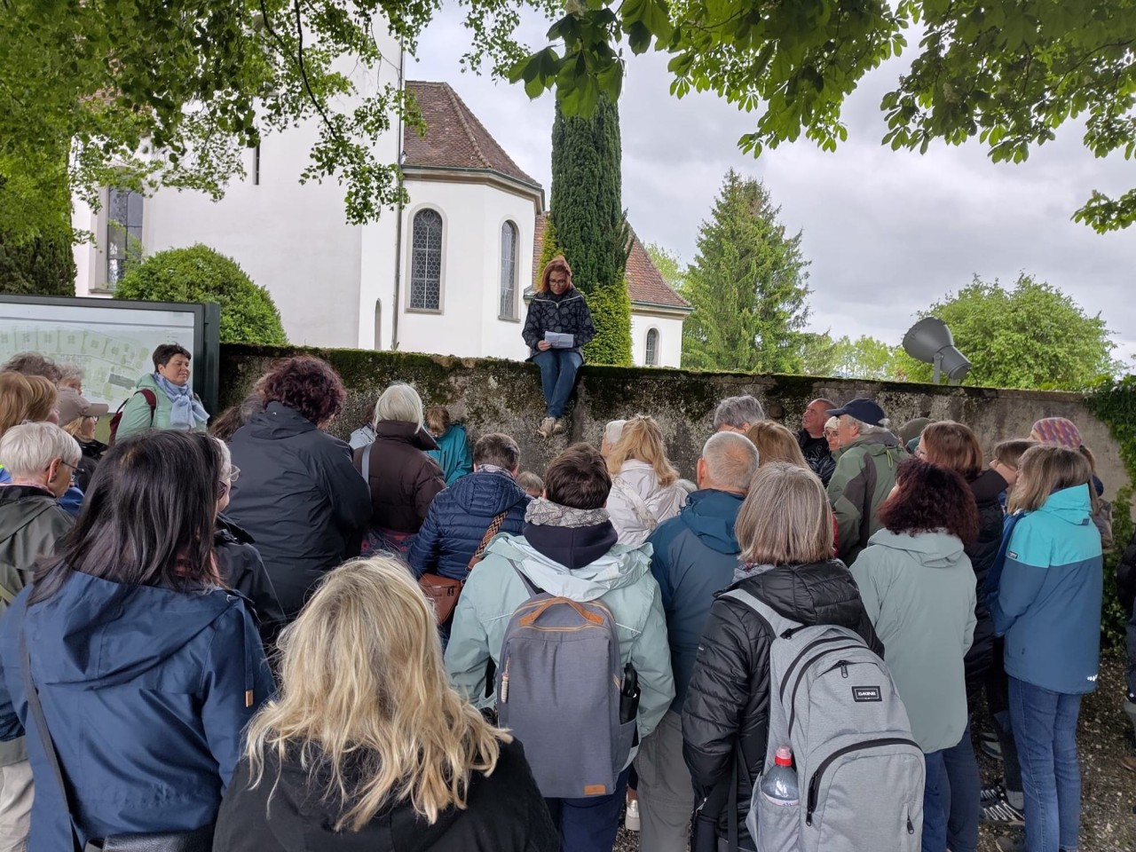 Frau sitzt vor einem Kloster auf einer Steinmauer und liest aus einem Buch. Vor ihr viele Menschen, die zuhören.