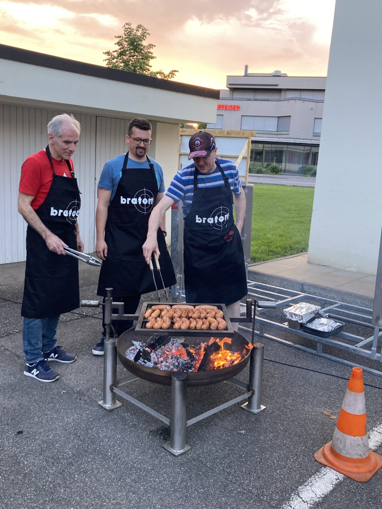 Drei Männer mit schwarzen Schürzen und Grillzangen beim Grillen im Freien.