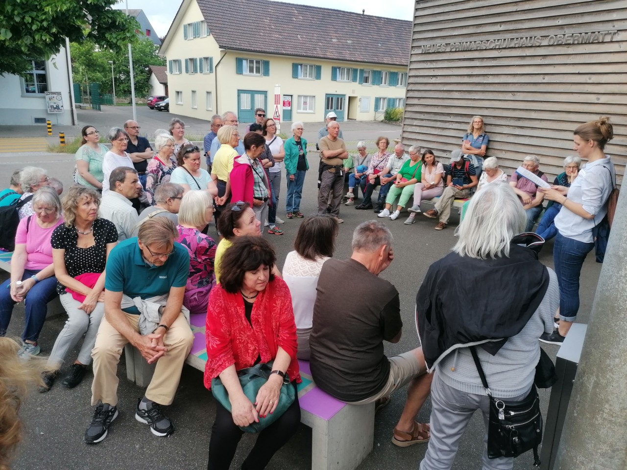 Personen sitzen draussen im Kreis und horen einer Frau zu, die stehend aus einem Buch liest.