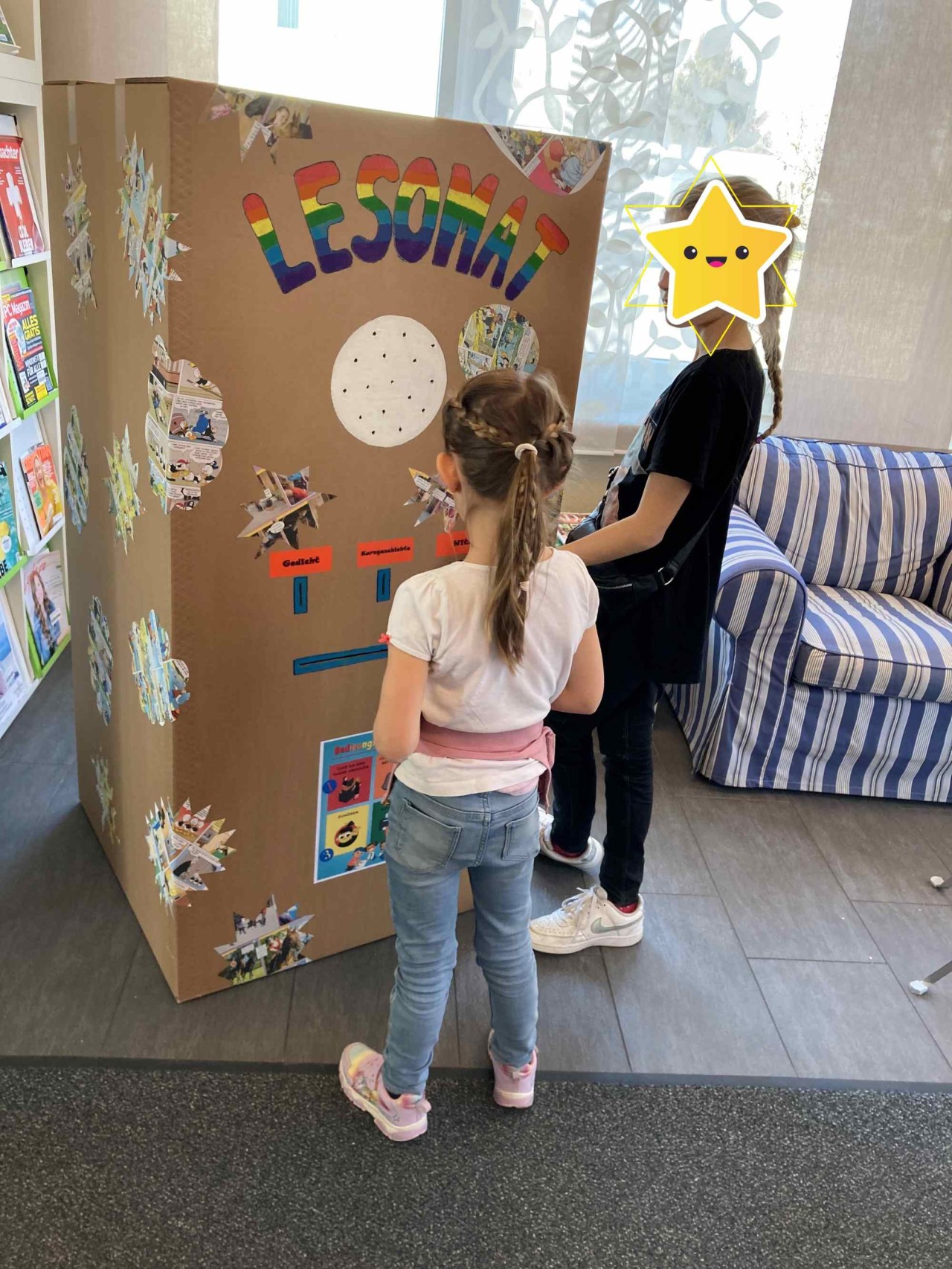 Zwei Mädchen stehen in einer Bibliothek vor einem grossen bunt beklebtem Karton mit der Aufschrift "Lesomat".