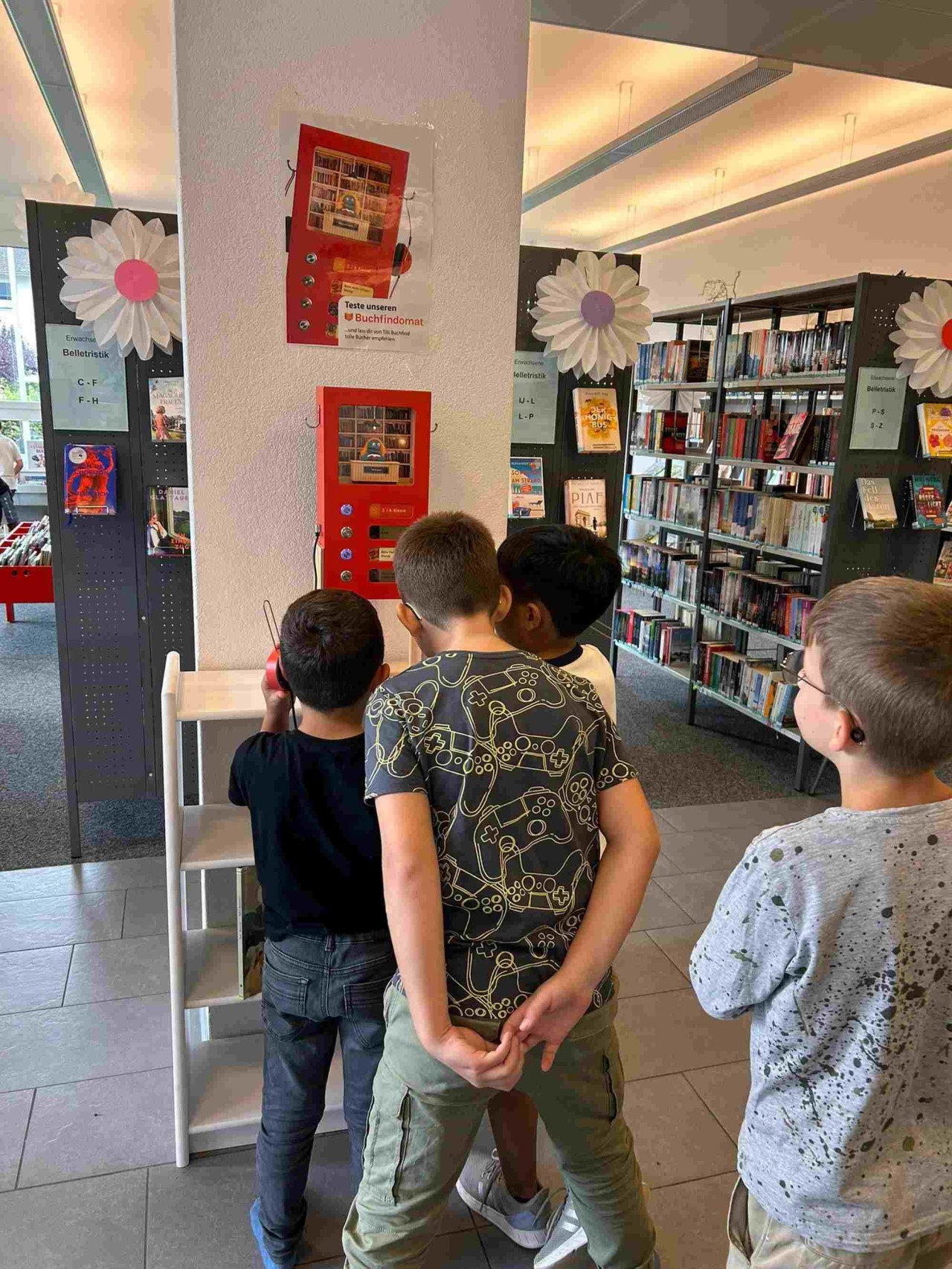 Vier Jungen stehen vor einem kleinen roten Automaten mit Knöpfen und Kopfhörern, in dem eine kleine Bibliothek eingerichtet ist.