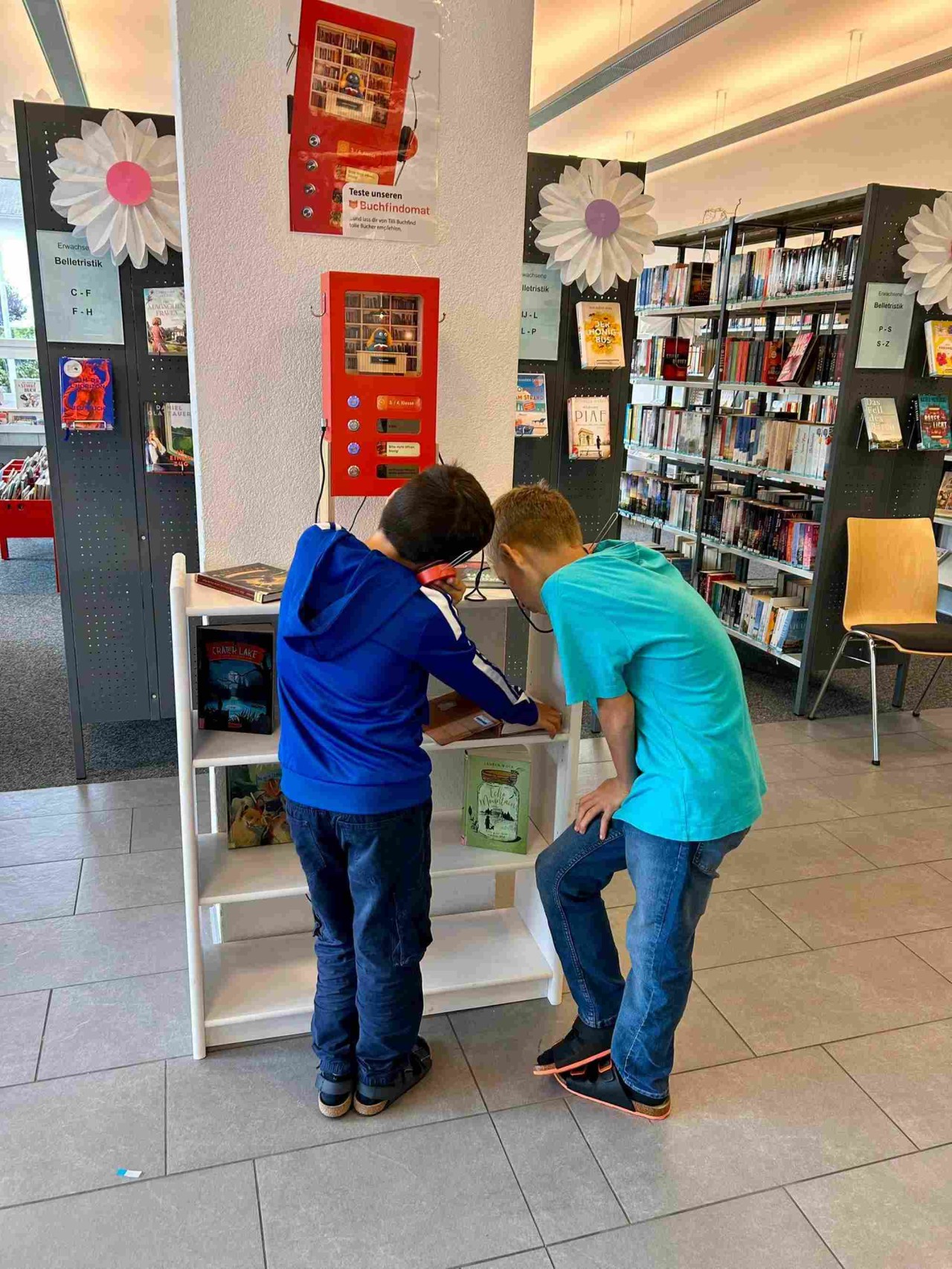 Zwei Jungen stehen vor einem kleinen roten Automaten mit Knöpfen und Kopfhörern, in dem eine kleine Bibliothek eingerichtet ist.