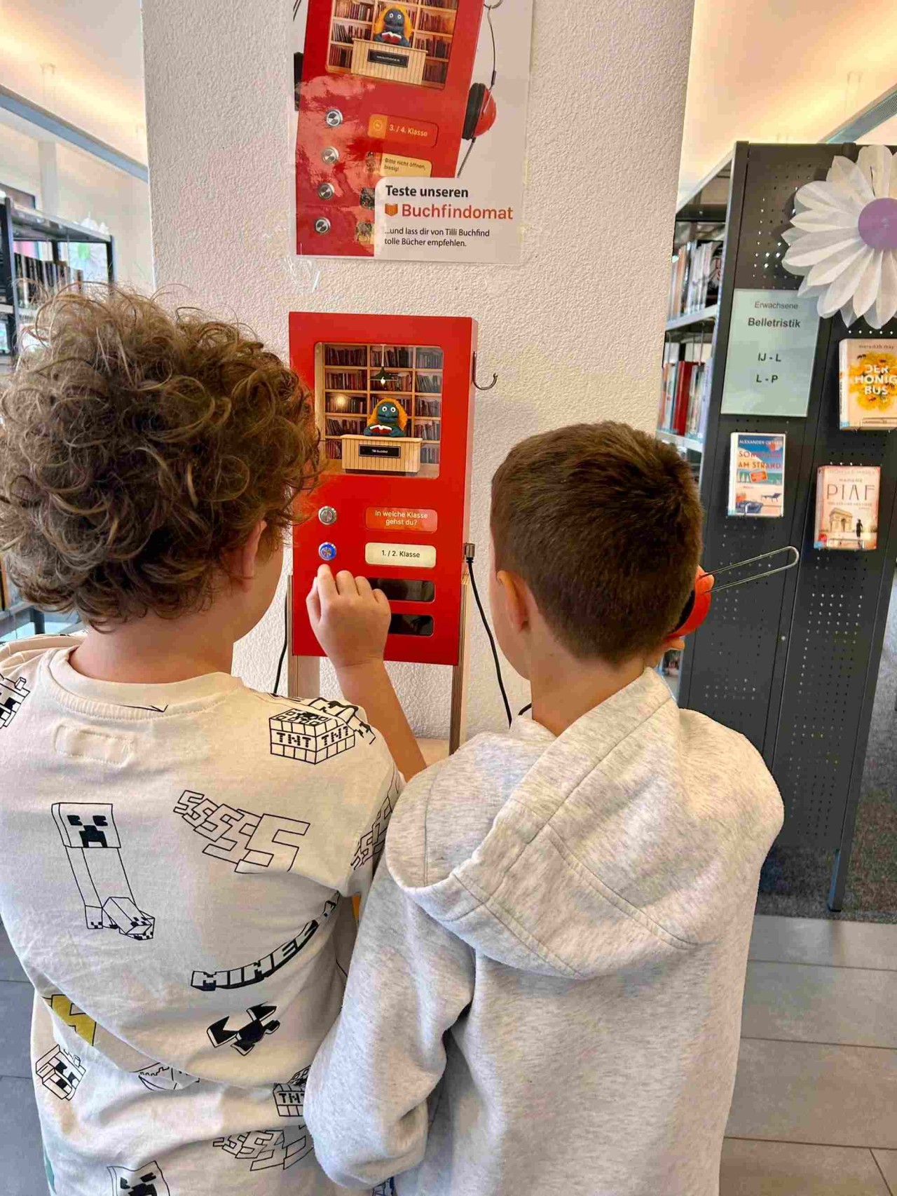 Zwei Jungen stehen vor einem kleinen roten Automaten mit Knöpfen und Kopfhörern, in dem eine kleine Bibliothek eingerichtet ist.