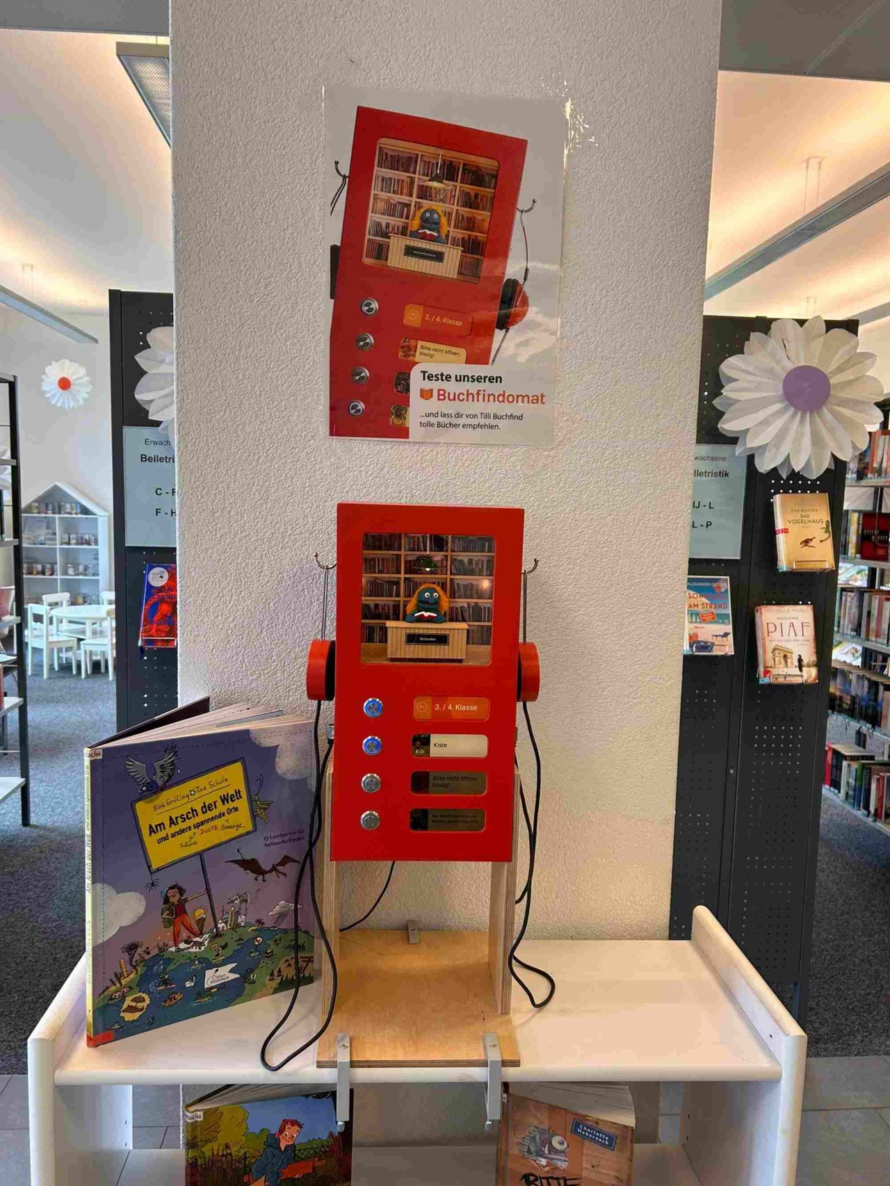 In einer Bibliothek steht ein kleiner roter Automat mit Knöpfen und Kopfhörern, in dem eine kleine Bibliothek mit sprechender Figur eingerichtet ist.
